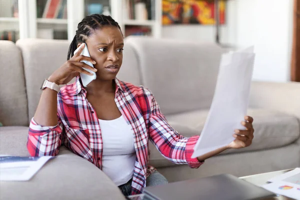 Irritated angry young black woman in casual entrepreneur sitting on floor at home, checking papers invoices, have phone call with assistant or business partner, copy space