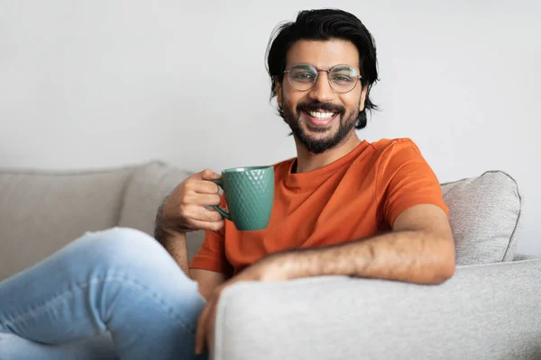 ガラスの明るい若い中東の男とコーヒーを飲みながらソファの上に座って リビングルームのインテリアでドリンクや無料の時間だけを楽しんでいます お茶を飲みながら家で休憩 — ストック写真