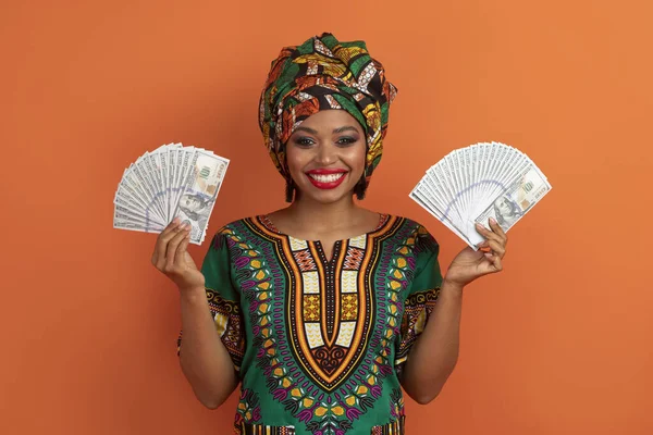伝統的なアフリカの衣類を身に着けている幸せな陽気な魅力的な若い黒人女性は 現金ドル紙幣と笑顔 オレンジスタジオの背景を保持し 賞金大きな宝くじを獲得しました 逃げろ賭けを — ストック写真