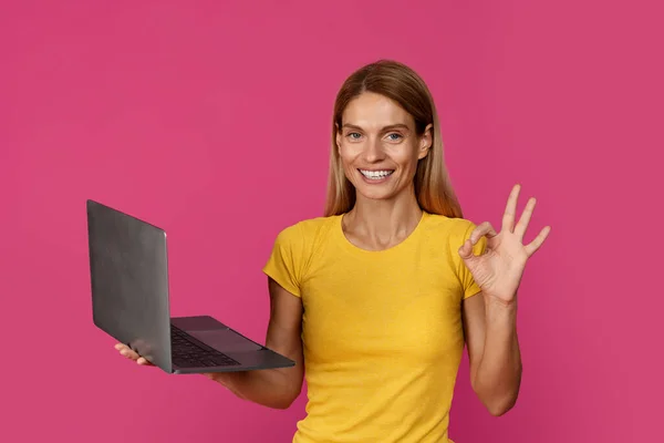 ラップトップを使用して幸せな中世ヨーロッパのブロンドの女性は ピンクのスタジオの背景に孤立した手でOkのサインを表示します ライフスタイル デバイスの承認 — ストック写真