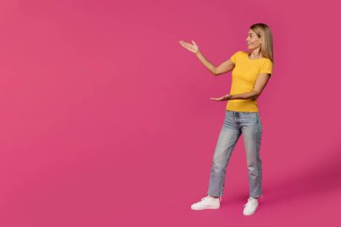 Şok olmuş orta yaşlı Avrupalı sarışın kadın ellerini pembe stüdyo arka planında tamamen izole edilmiş boş bir yere doğrultuyor. Büyük bir satış, sürpriz, harika haberler, insanların duyguları ve sunumu