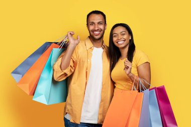 Neşeli bin yıllık Afro-Amerikan erkek ve kadın birçok alışveriş çantası bulundurur, sarı stüdyo arka planında izole edilmiş alışverişin tadını çıkarırlar. Alıcılara, alışverişe ve modaya özel satış