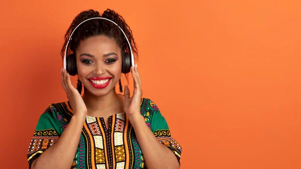 stock image Closeup of positive happy smiling attractive young black woman wearing bright makeup and african traditional shirt using modern wireless headphones, listening to music, orange background, copy space