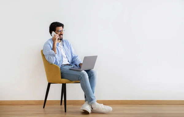 身穿休闲装的快乐的年轻穆斯林小伙子 戴着眼镜坐在扶手椅上 手持笔记本电脑 在灰色墙上的房间里打电话 远程通信 生活方式 自由职业 在家做生意 — 图库照片