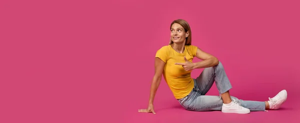 前向きな中世ヨーロッパのブロンドの女性が床に座っているピンクのスタジオの背景 パノラマに隔離された空のスペースで指を指しています 推薦広告と提供 人間の感情 — ストック写真