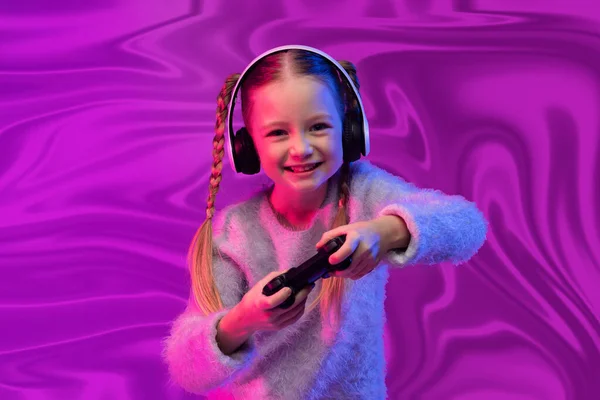 快乐快乐的笑着的小女孩金发碧眼的学龄女孩在抽象的紫外线波浪形背景下玩电子游戏 用无线耳机和手握操纵杆 儿童的游戏成瘾 — 图库照片