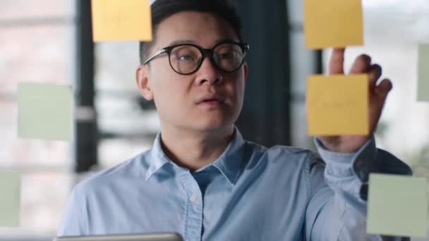 深刻な中年アジア人男性チェックスケジュールでデジタルタブレット 読書ノートオンステッカー 近くに立ってガラス壁で現代オフィス クローズアップ肖像画 — ストック動画