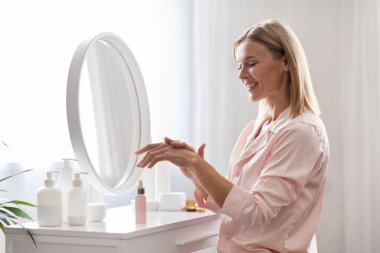 Cilt nemlendirici. Gülümseyen orta yaşlı kadın, evde el kremi sürüyor, güzel olgun bayan besleyici serum kullanıyor, yatak odasında giyinme masasında otururken günlük cilt bakımı rutini yapıyor.