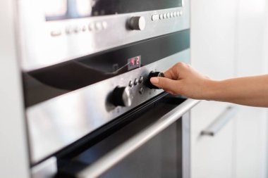 Mutfakta elektrikli fırın kullanan tanınmayan bir kadın, sıcaklığı eliyle ayarlayan, düğmeye basan genç bir ev hanımı, evde yemek hazırlarken yakıcı modunu seçen, yakın çekim yapan bir kadın.