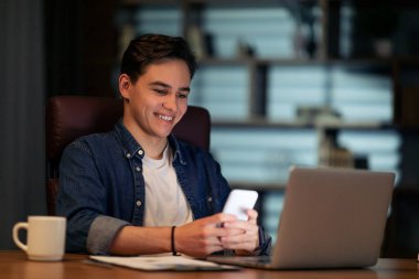 İş iletişimi ve modern teknolojiler. Neşeli genç girişimci karanlık boş ofiste çalışma masasında oturuyor, dizüstü bilgisayar ve akıllı telefon kullanıyor, e-postaları kontrol ediyor, boşlukları kopyalıyor.