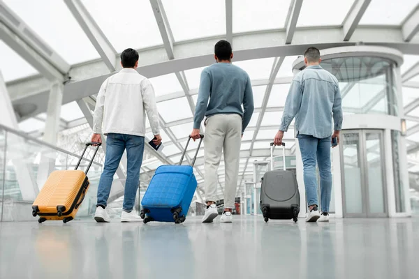 Cheap Travel Tickets. Rear View Of Three Male Friends Group Walking With Suitcases Posing Back To Camera In Airport Terminal Inside. Full Length Shot Of Unrecognizable Passengers Guys