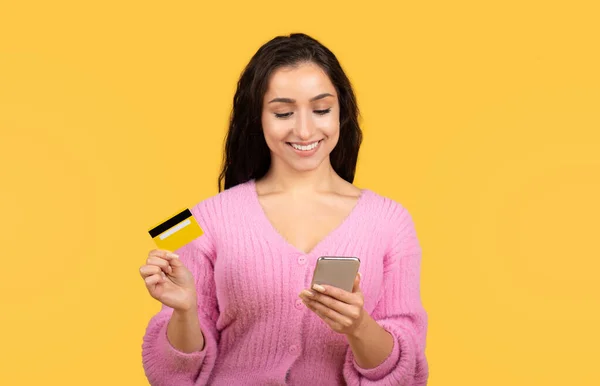 クレジットカードやスマートフォンを搭載した笑顔の千年アラブ女性は 黄色のスタジオの背景に隔離されたオンラインショッピングをお楽しみください 巨大な販売 キャッシュバックのための金融アプリ キャッシュバック 広告とオファー — ストック写真