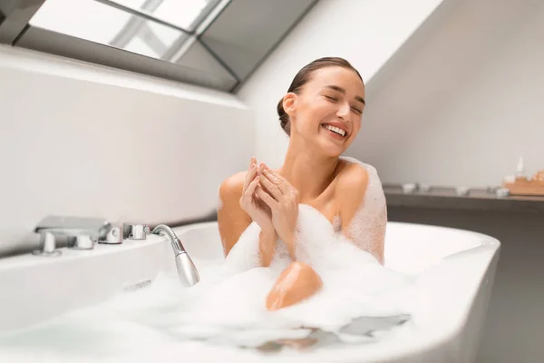 泡でお風呂に入る陽気な女性自宅で近代的なバスルームで目を閉じて洗濯体をリラックスさせます ソープスッドで覆われた魅力的な若い女性の入浴ポーズ — ストック写真