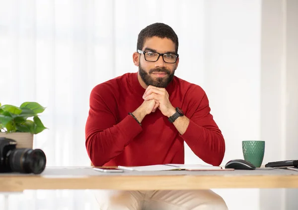 stock image Happy smiling handsome hispanic young man wearing stylish casual outfit and eyeglasses photographer posing at workplace, guy sitting at desk with computer, gadgets and notepad, drinking coffee