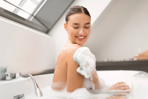 美と衛生 スポンジで幸せな女性こすり体は自宅でお風呂に泡で覆われて座っています 女性はリラックスして自分自身の世話を入浴 ウェルネス スパ化粧品 — ストック写真