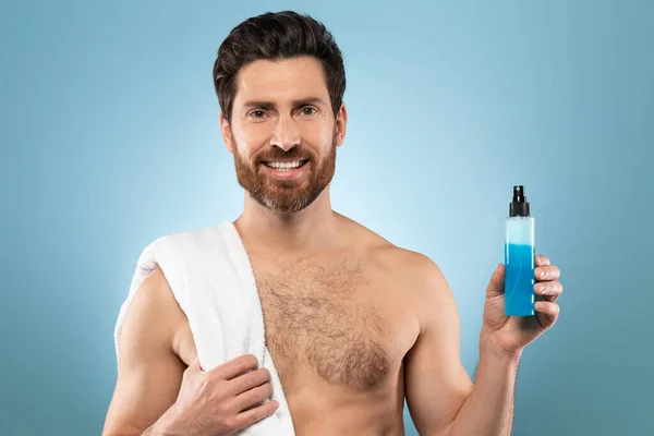 ハンサムな中年男性とともにタオルで肩の上にボトルを見せます青い化粧品液体 青いスタジオの背景の上にポーズと笑顔でカメラ フリースペース — ストック写真