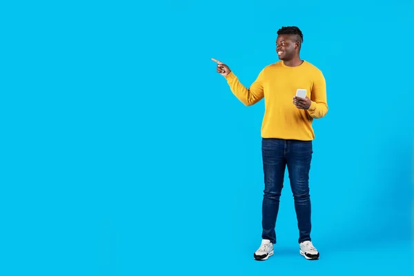 看看这个黑人手持智能手机 带着手指在复印空间边指指指点点 站在蓝色背景下笑着为年轻的非裔美国男性做广告 — 图库照片