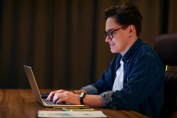 Remote job, freelance. Side view of positive young guy wearing eyeglasses and casual outfit sitting at desk late at night at home, typing on laptop keyboard, independent contractor working late