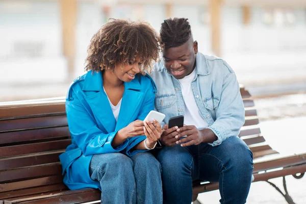 ベンチの屋外に座っている間にスマートフォンを使用して陽気な黒のカップル 幸せな若いアフリカ系アメリカ人男性と女性がWifiを共有するか 彼らの携帯電話で写真を見て 閉鎖ショット — ストック写真