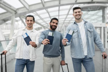 Ucuz Bilet Teklifi. Modern havalimanında seyahat bavullarıyla kameraya gülümseyen neşeli erkek turist grubu biniş kartlarını gösteriyor. Pasaportlara Seçici Odaklanma
