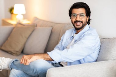 Neşeli, yakışıklı, orta doğulu, rahat giyinen ve gözlüklü genç adam kanepede oturur, dinlenir, oturma odasının içinde boş vakit geçirir. Yaşam tarzı, rahatlık, hafta sonu evde yalnızlık