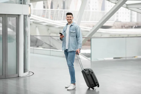 旅行申请 快乐旅行者的家伙使用手机微笑相机站在机场室内与手提箱 男性乘客通过电话发短信报站 自由空间 — 图库照片