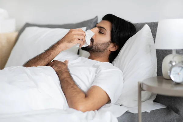 身穿白色T恤的可悲的中东年轻人躺在床上 用餐巾纸吹鼻涕 在卧室里得了流感和感冒 在家里治疗 — 图库照片