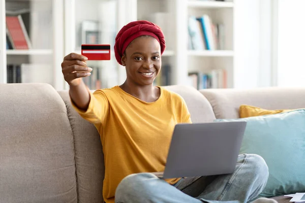 Happy smiling beautiful young black woman in red turban using laptop, showing bank card at camera, sitting on couch at home, order food online, paying for goods and services on Internet, copy space