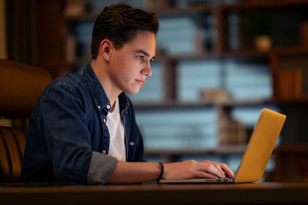 Concentrated handsome young guy developer typing on modern pc laptop keyboard, coding at dark office, staying late at night, have deadline, employee overworking, side view, copy space