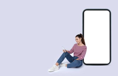 Gülümseyen hoş, şık, zarif Latin bir kadın yerde oturuyor, beyaz ekranlı büyük bir cep telefonuna yaslanıyor, akıllı telefon kullanıyor, model yapıyor, gri stüdyo arka planında izole edilmiş, boşluğu kopyalıyor.