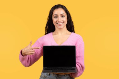 Bin yıllık neşeli Arap kadın işaret parmağı dizüstü bilgisayarda ve sarı stüdyo arka planında boş ekran var. Sunum, tavsiye, web sitesi, blog, uygulama, harika haberler, reklam ve teklif