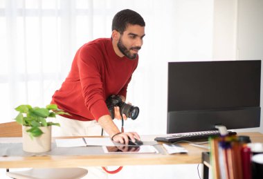 Yaratıcı bir iş. Yakışıklı sakallı genç adam, günlük kıyafetli profesyonel fotoğrafçı, fotoğraf stüdyosunda çalışıyor, dijital kamera ve bilgisayar kullanıyor, fotoğraf çekimini kontrol ediyor, uzayı kopyalıyor.
