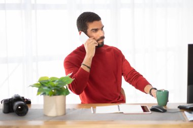 Yakışıklı İspanyol asıllı sıradan fotoğrafçı bilgisayarlı, DSRL kameralı, iş yerindeki dijital tabletli, müşterisiyle telefon görüşmesi yapan, mekanı kopyalayan.