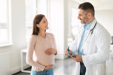 Doktor randevusu. Muayene sırasında terapistle konuşan mutlu ve hamile bir kadın. Karnını kucaklayan ve gülümseyen bir kadın.