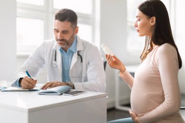 Genç hamile bir kadın için reçete yazan erkek doktor, elinde ilaç şişesi olan kadın, reçeteyi açıklayan terapist ve hamile olan genç anneye vitamin ya da ilaç veren kadın.