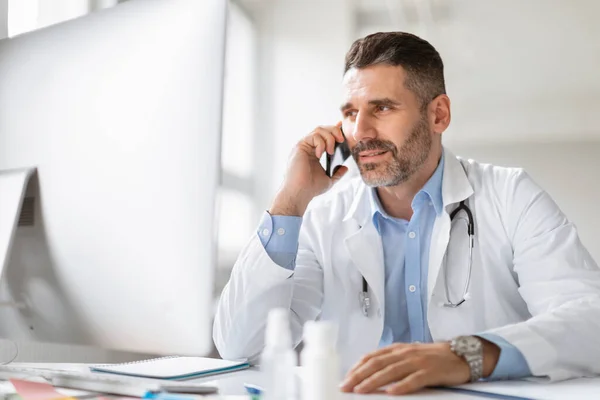 快乐的男医生 与病人进行电话交谈 分享良好的检查结果或提供专业的医疗咨询 坐在诊所的桌子旁 — 图库照片