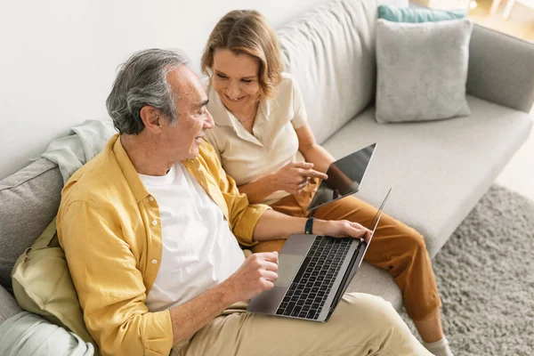 ラップトップコンピュータとデジタルタブレットを使用して現代の高齢者のカップルは 自宅でソファに座って話しています ガジェットを持っている配偶者 インターネットを閲覧 技術コンセプト — ストック写真