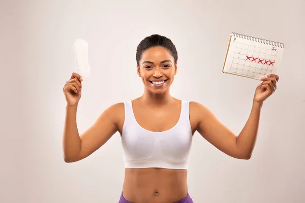 Happy cheerful young black woman demonstrates periods calendar and sanitary napkin, isolated on grey studio background. Women healthcare, menstruation, menstrual cycle concept