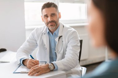 Genel pratisyen doktor randevusu. Kadın hastasıyla konuşan, teşhis ve tedaviyi tartışan kendinden emin bir erkek doktorun portresi. Ofisinde kadınlara danışmanlık yapan bir uzman.