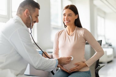 Tıbbi servis konsepti. Erkek doktor hamile kadınların karnını steteskop kullanarak muayene ediyor. Ofisteki bebeklerin kalp atışlarını dinliyor. Bekleyen kadın doktora bakıyor ve gülümsüyor.