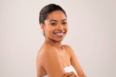 Mutlu, yarı çıplak, parlak tenli, kameraya gülümseyen, gri stüdyo arka planında poz veren, mükemmel cildini, fotokopi alanını ve afişini gösteren siyah bir kadın. SPA konsepti