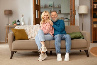 Mutlu evlilikler. Neşeli olgun eşler modern oturma odasındaki koltukta oturmuş kameraya gülümsüyor ve evde poz veriyor. Yaşlılıkta Romantik İlişki ve Aşk