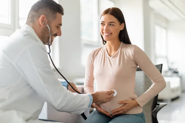 医疗服务概念 男医生用听诊器检查孕妇的腹部 在办公室里听婴儿的心跳 期待中的女士看着医生 微笑着 — 图库照片