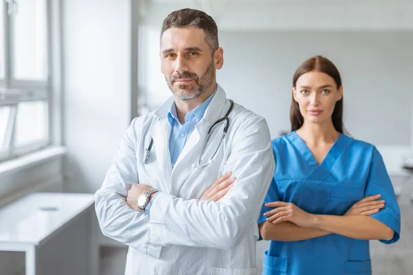 Medical services concept. Confident doctor male and female colleagues wearing coats, posing with folded arms and looking at camera, standing in modern light medical office