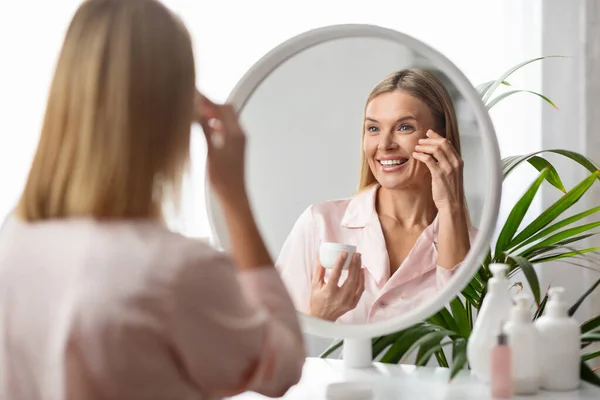 美丽的中年妇女照镜子 应用润肤眼霜 快乐的微笑成熟的女性在家做抗衰老的美容治疗 享受自我照顾 有选择的关注 — 图库照片