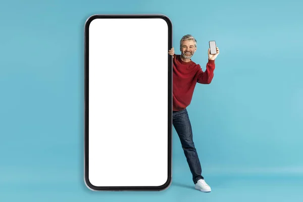 有魅力 休闲的男人 站在白色空白屏幕的大型智能手机后面 展示手机和微笑 推荐手机应用 蓝色背景 — 图库照片