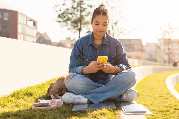 黑人女大学生使用手机 在社交媒体上发短信 坐在草坪上 复制空间 拿着智能手机的女学生人和小玩意 — 图库照片