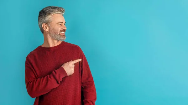 Check this out. Handsome grey-haired bearded middle aged man in red pointing at copy space for advertisement or text and smiling, isolated on blue studio background, panorama