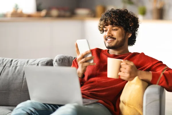 Bonito Indiano Guy Relaxante Casa Com Smartphone Laptop Coffee Sorrindo — Fotografia de Stock