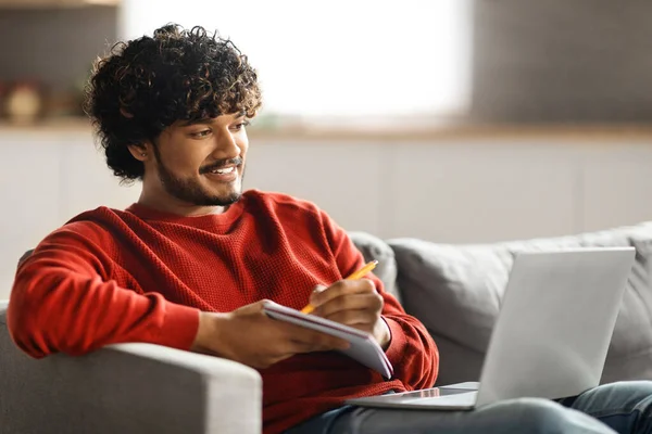 Online Education. Handsome young indian man using laptop and taking notes while sitting on couch at home, smiling eastern guy watching tutorial, lecture or webinar, looking at computer screen
